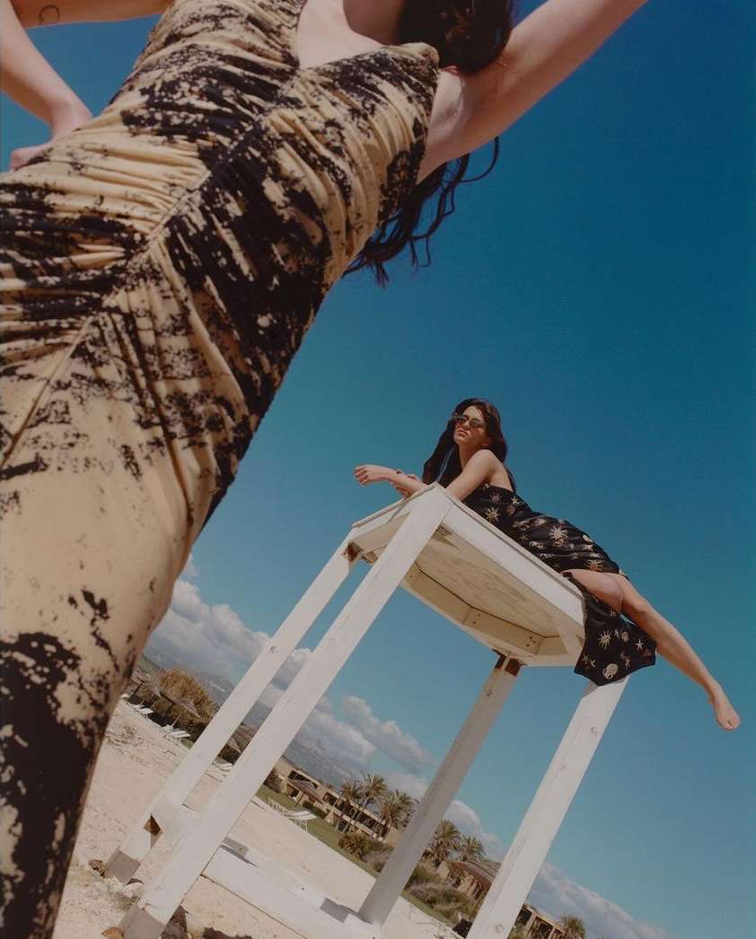 Two women in dresses pose outdoors on a sunny day. One woman stands in the foreground, the other sits on a white structure.