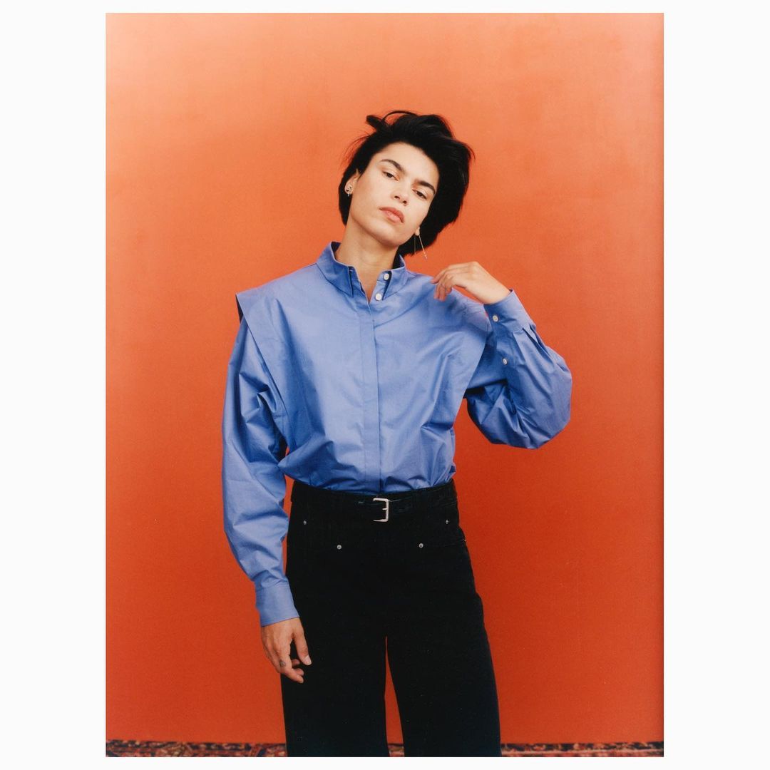 An adult female poses indoors wearing a blue dress shirt, black pants with a belt, and jewelry in front of an orange background.