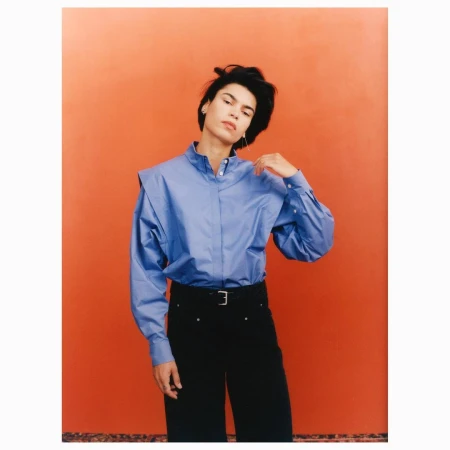 An adult female poses indoors wearing a blue dress shirt, black pants with a belt, and jewelry in front of an orange background.