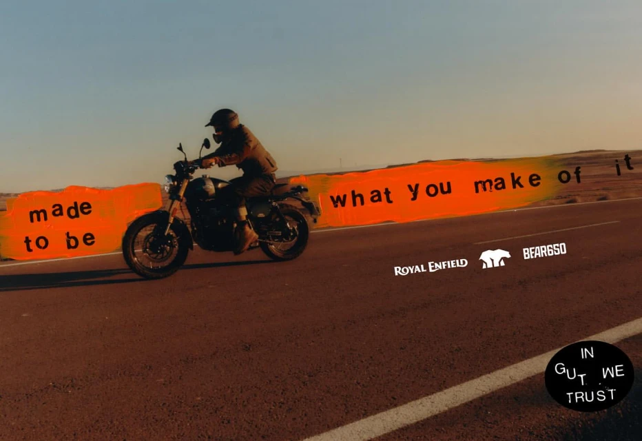Person riding a Royal Enfield motorcycle on a desert highway with 'Made to be what you make of it' text, under a bright sky.