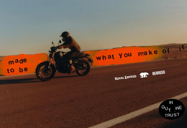 Person riding a Royal Enfield motorcycle on a desert highway with 'Made to be what you make of it' text, under a bright sky.