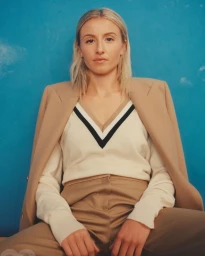 Portrait of a blonde woman wearing a tan blazer and a v-neck knit sweater against a bright blue wall