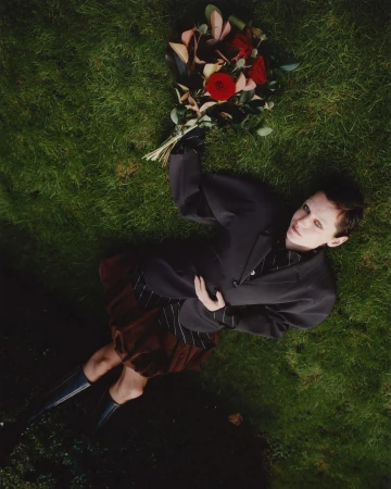 Person lying on green grass holding a bouquet of red and pink roses, wearing a dark blazer and brown skirt.