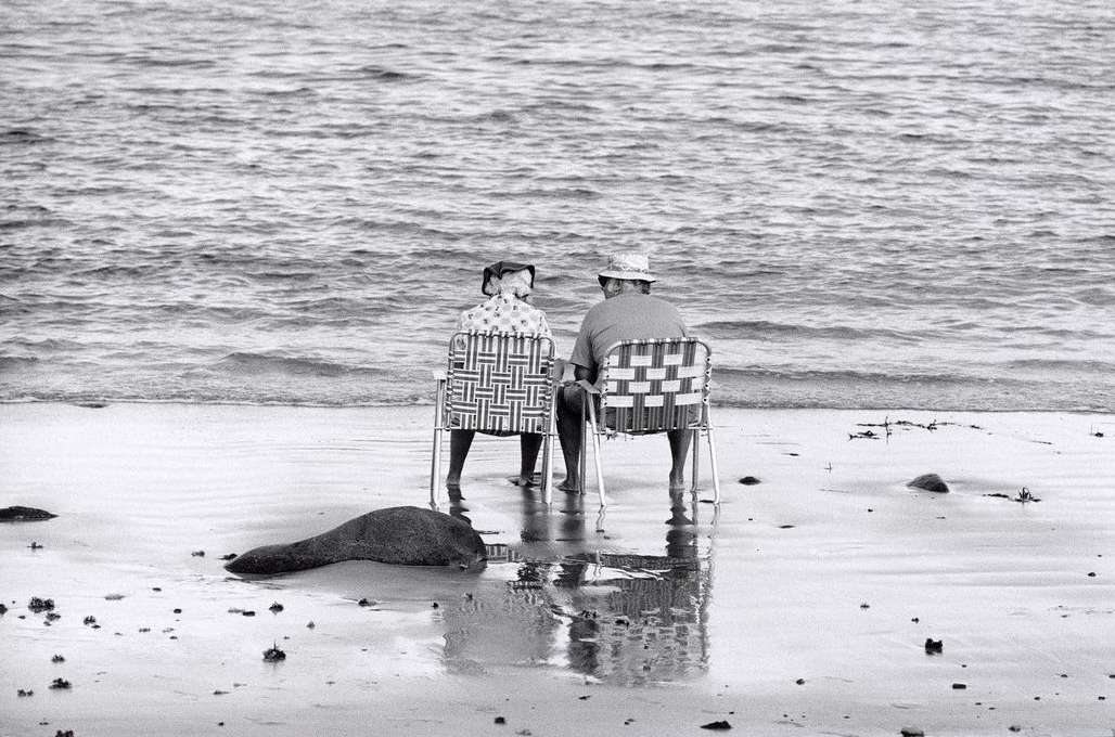 Elderly couple sits in woven chairs on a sandy beach next to the ocean. Black and white photography featuring seascape and relaxation.
