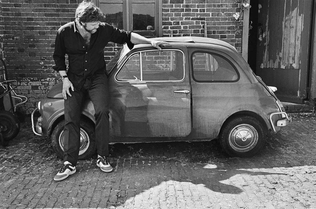 Adult male in a black shirt leans against a vintage compact car on a brick road, with a brick building visible in the background.