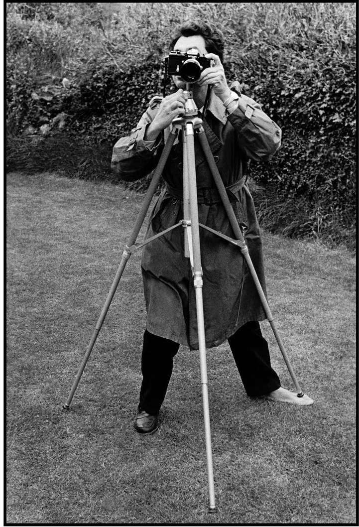 Photographer with vintage camera on tripod in grassy outdoor setting, capturing the art of photography in black and white.