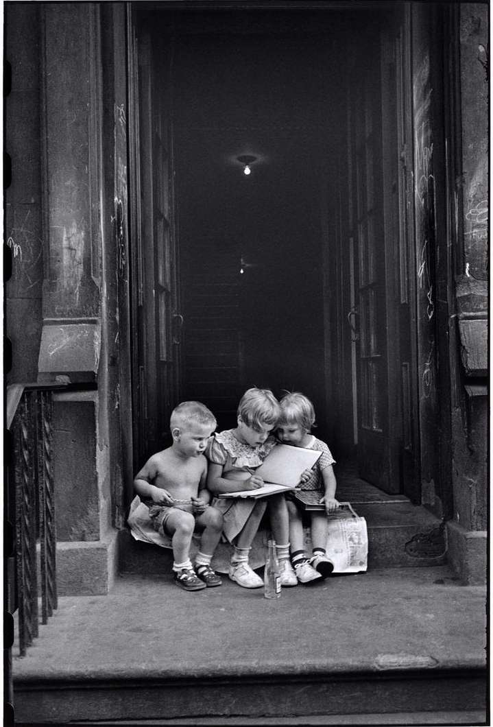 Three young children sit on a stoop together, looking at papers in a black and white photograph taken outdoors in daylight.