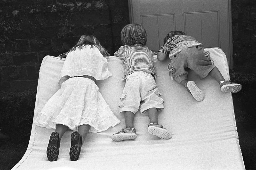 Three children lying on a white surface outdoors, captured in black and white, with a focus on their backs and the details of their clothing.