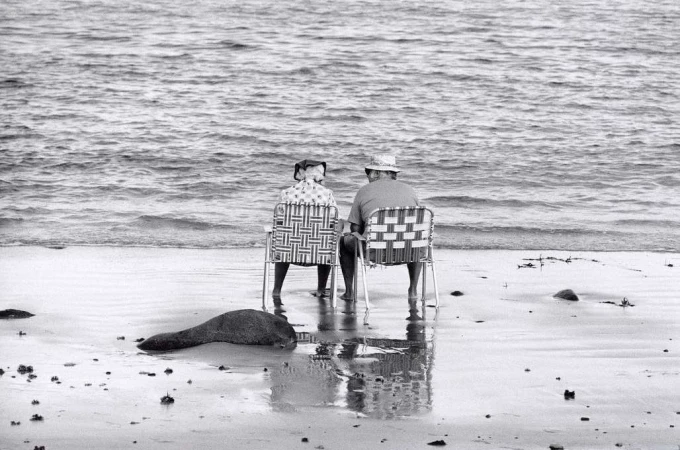 Two elderly people seated in folding beach chairs looking out at the ocean on a sandy beach.