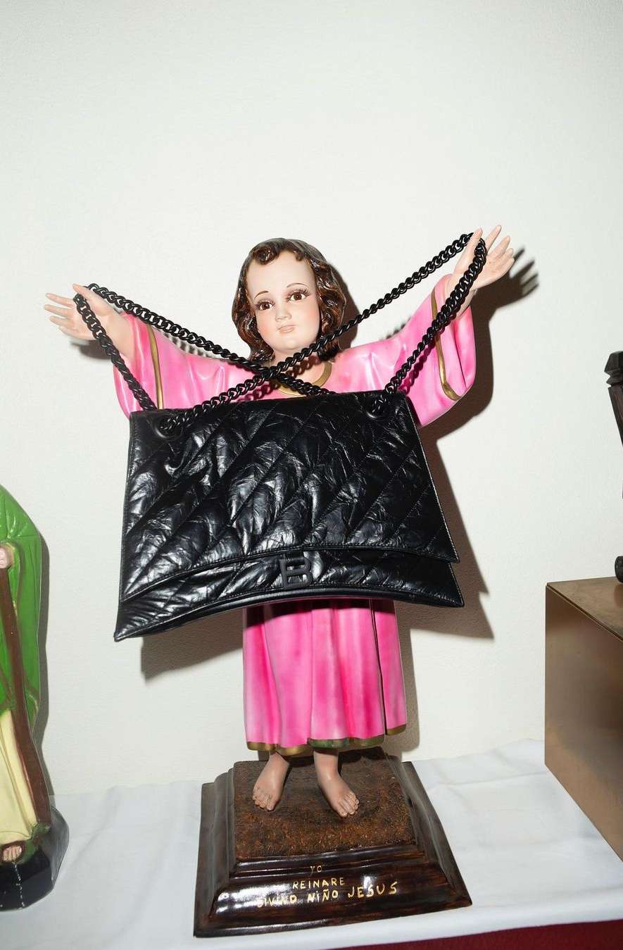 Black quilted handbag draped over a pink-robed religious statue on a brown base against a white background in an indoor setting.