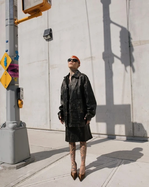 Stylish woman in black leather jacket and skirt standing on city sidewalk near traffic light on sunny day, posing with confidence.