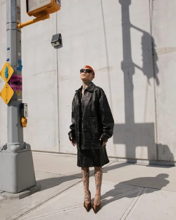 Stylish woman in black leather jacket and skirt standing on city sidewalk near traffic light on sunny day, posing with confidence.