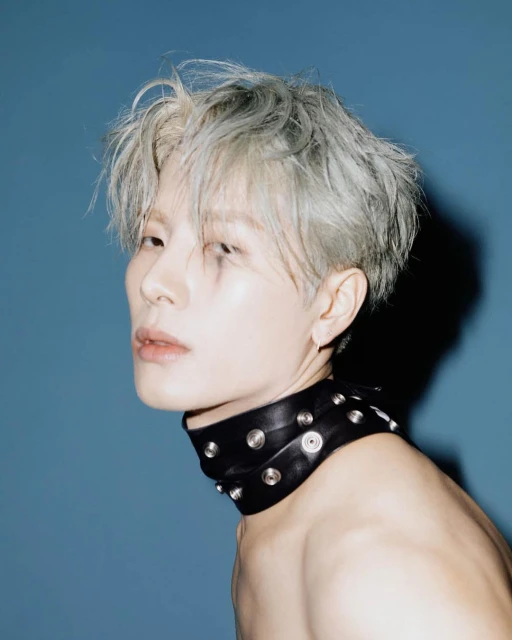 Portrait of a young adult with grey hair, wearing a studded leather choker necklace, against a blue background in a photo studio.