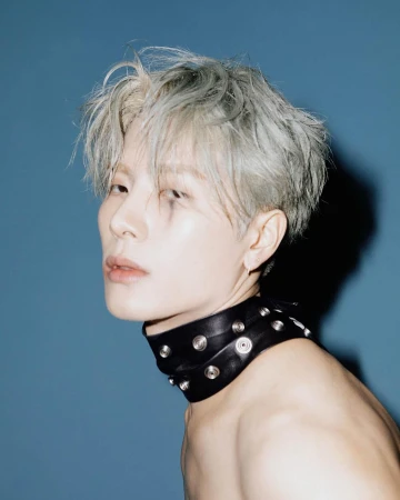 Portrait of a young adult with grey hair, wearing a studded leather choker necklace, against a blue background in a photo studio.