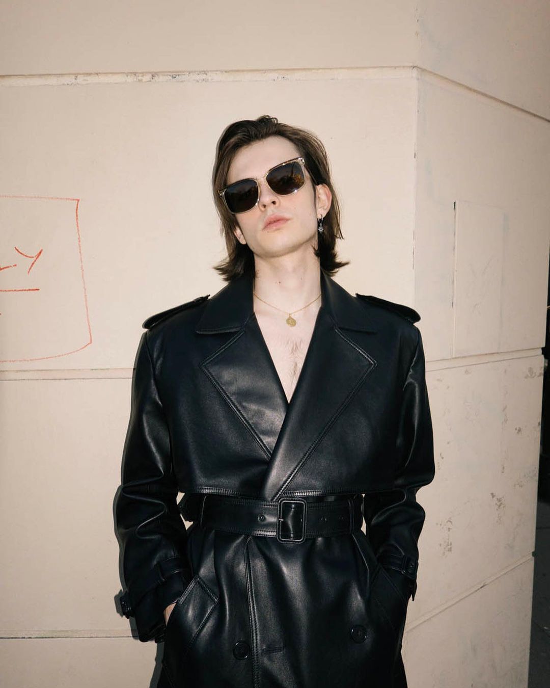 Man with dark hair wearing black sunglasses and a black leather trench coat standing against a beige wall