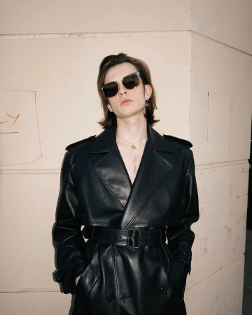 Man with dark hair wearing black sunglasses and a black leather trench coat standing against a beige wall