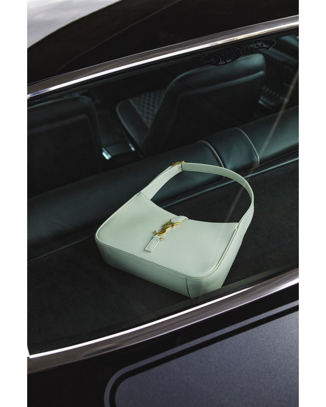 Light blue leather shoulder bag with gold YSL logo hardware displayed inside a dark vehicle with black leather interior.