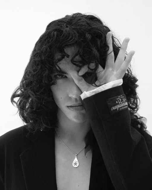 Portrait of a young adult with curly dark hair wearing a black blazer and a teardrop pendant necklace with a star against a white background.