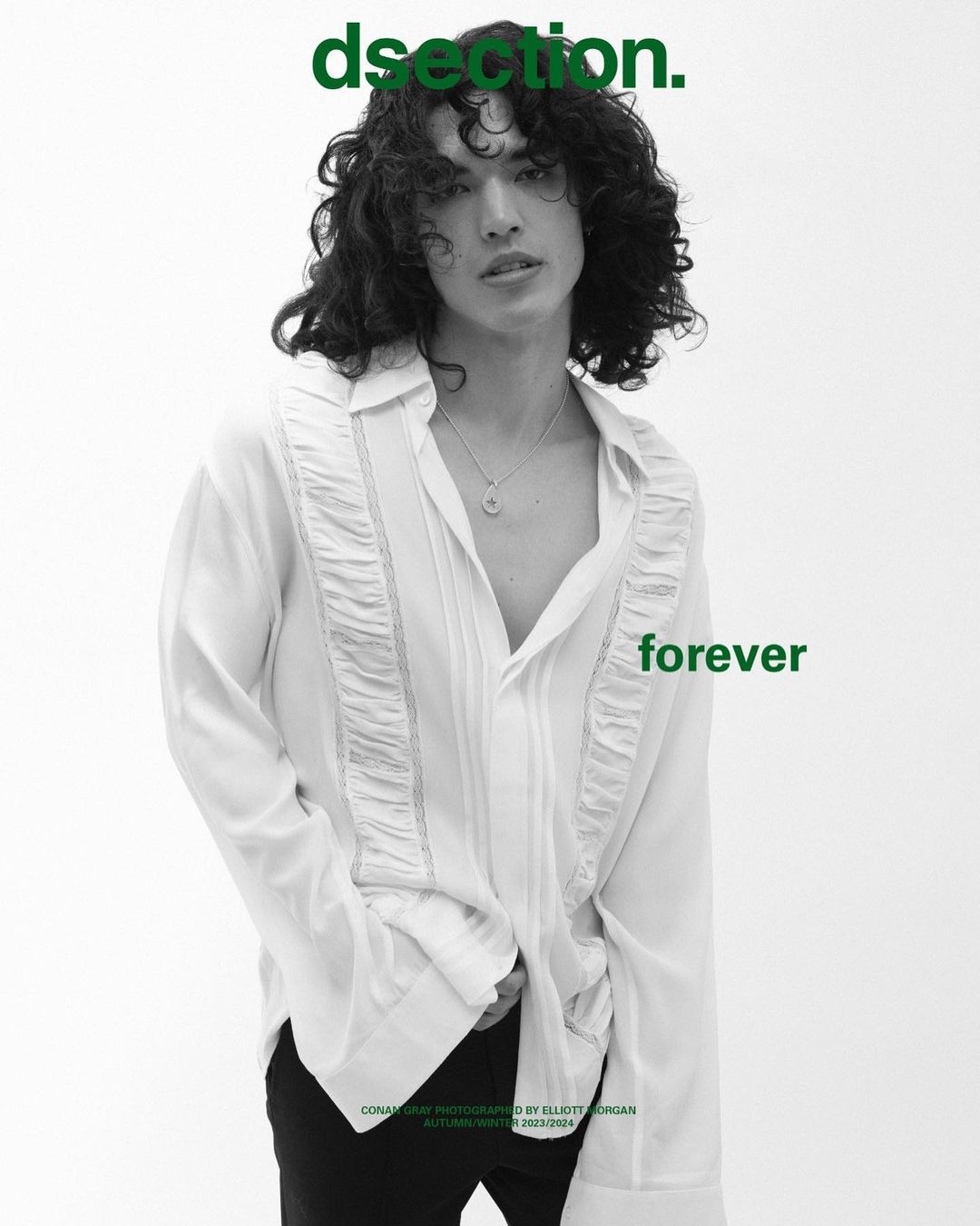 Young adult male with dark curly hair poses in an open white shirt and black pants against a white backdrop in a studio setting.