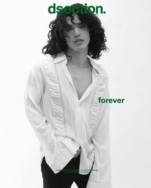 Young adult male with dark curly hair poses in an open white shirt and black pants against a white backdrop in a studio setting.