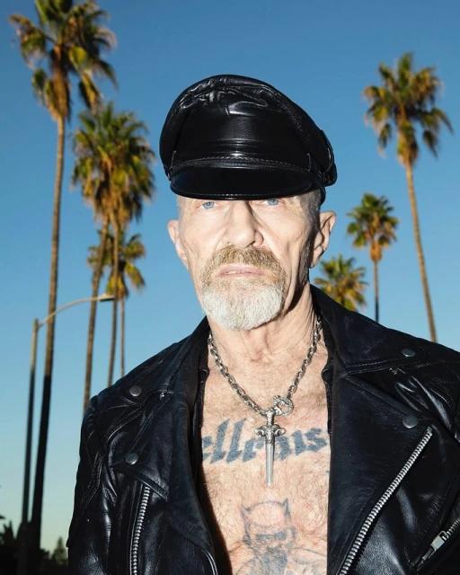 Portrait of a mature man in a leather cap and jacket posing outdoors with palm trees against a blue sky in the background.