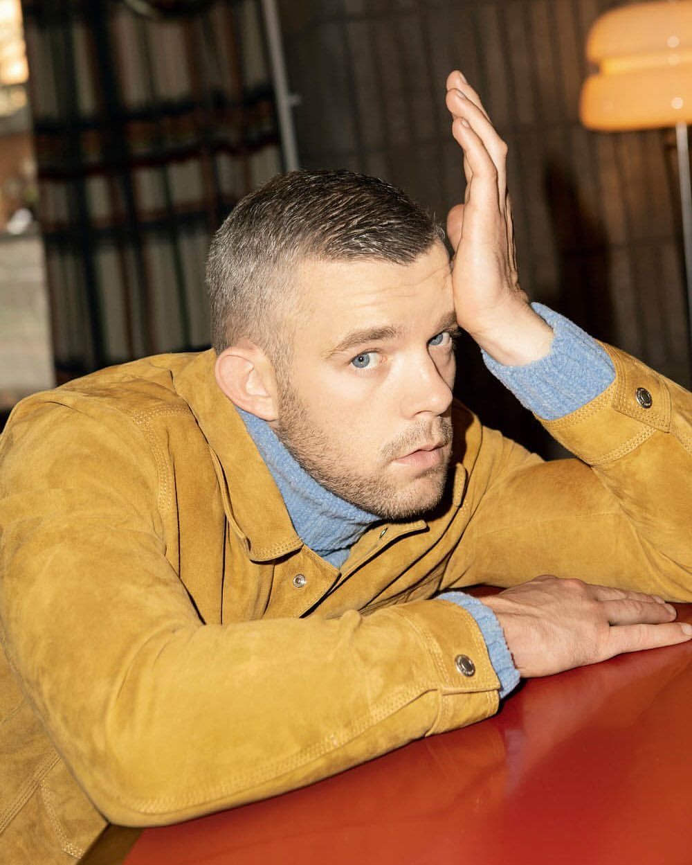 Indoor portrait of a short-haired male model in a yellow jacket resting his arm on a red table, gazing directly at the camera.