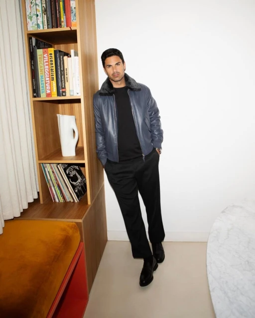 Adult male in a blue leather jacket poses indoors next to a wooden shelf with books and a white pitcher, creating a stylish portrait.
