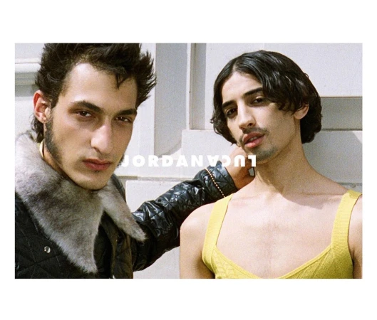 Two adult men pose indoors in a medium shot. One man wears a black jacket with fur collar, the other a yellow tank top against a white wall.