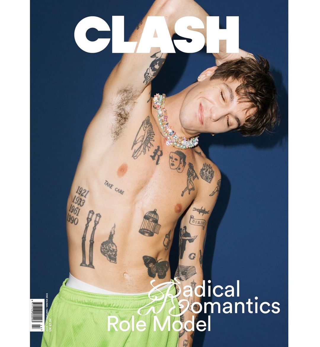 Shirtless man with tattoos and colorful necklace poses for a Clash magazine cover against a solid blue background. He is smiling.
