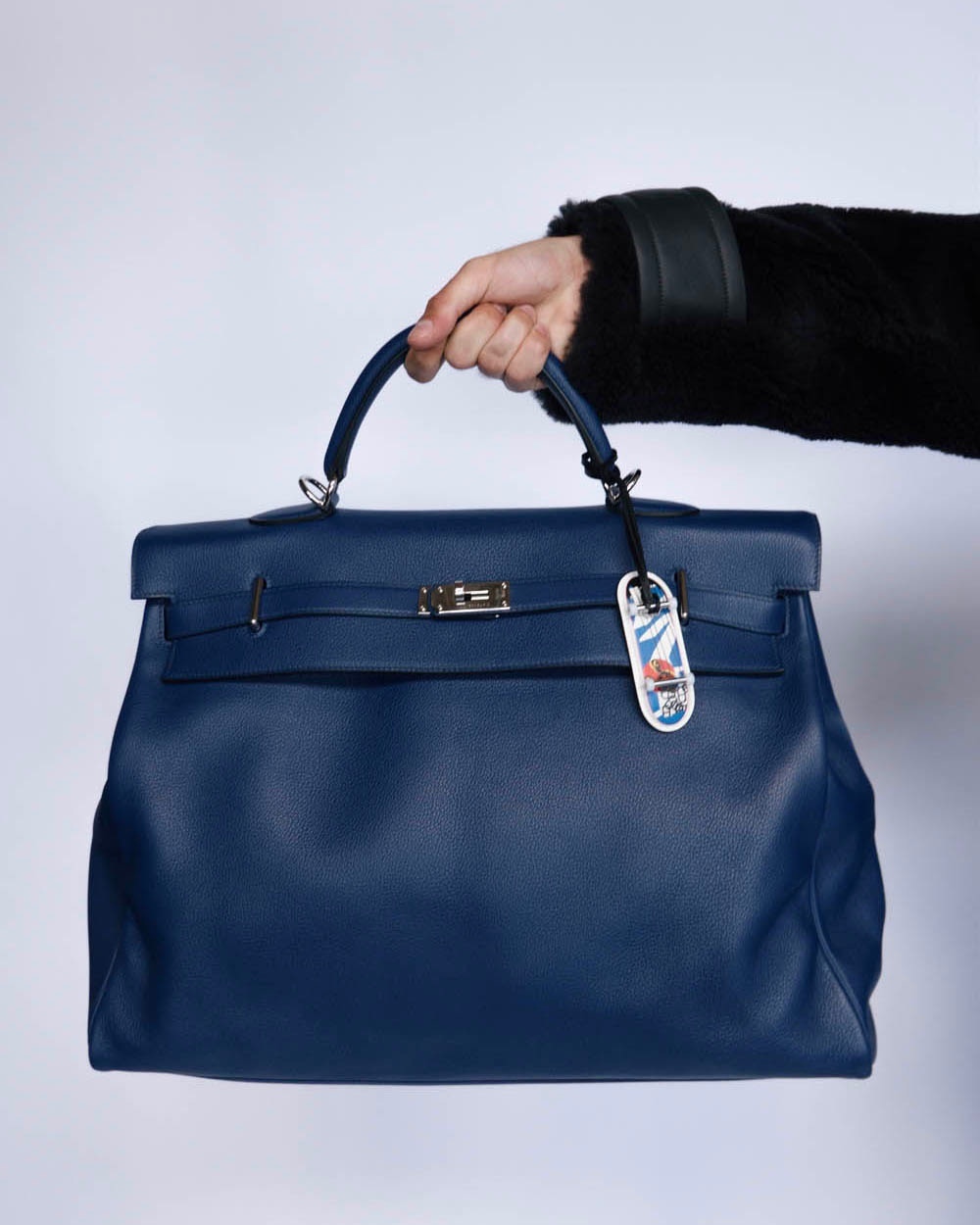Close-up of a person holding a blue leather Hermès Kelly bag, featuring silver hardware and a decorative tag against a white background.