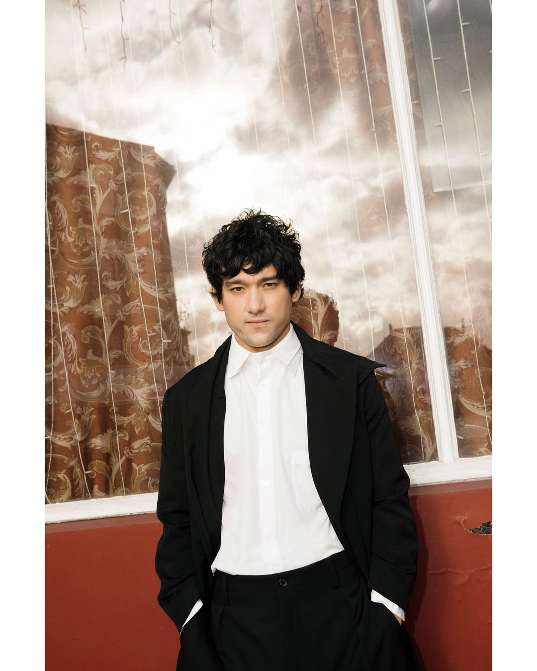 Portrait of an adult male wearing a black coat and white shirt, leaning against a wall in front of a large window with patterned curtains.