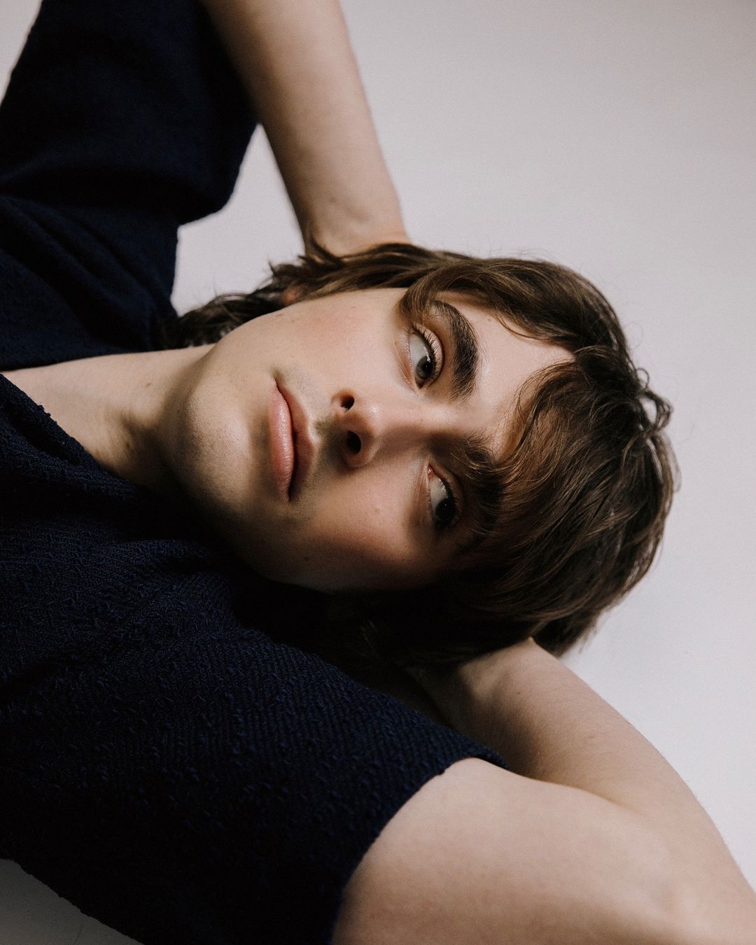 Portrait of a young adult male with brown hair, gazing slightly off-camera. He is lying down with arms behind his head against a plain background.