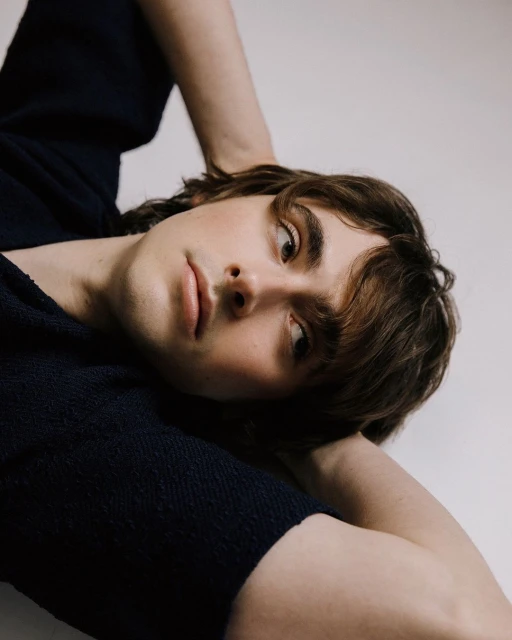 Portrait of a young adult male with brown hair, gazing slightly off-camera. He is lying down with arms behind his head against a plain background.