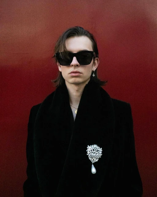 Close-up studio shot of a young adult male wearing sunglasses and a black velvet coat with a large pearl brooch, set against a dark red background.