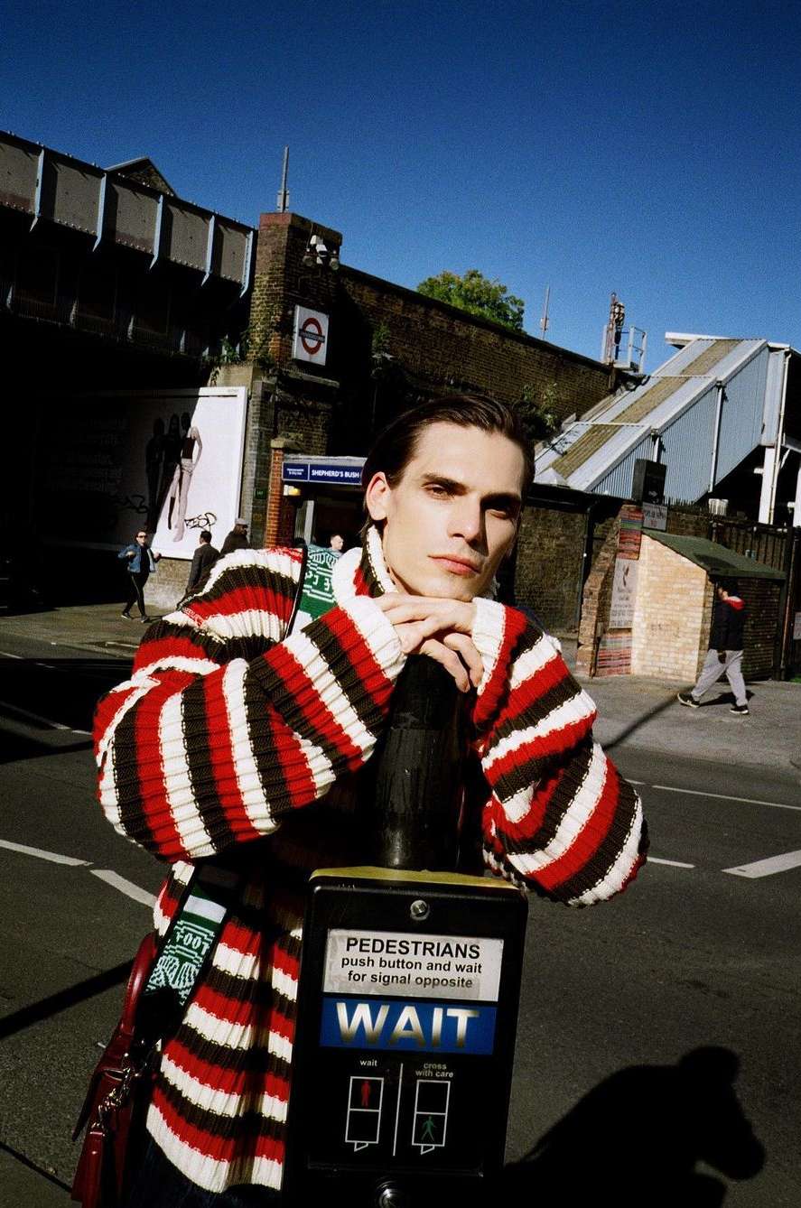 A young adult poses against an urban background. He wears a striped sweater while leaning on a crosswalk signal pole.