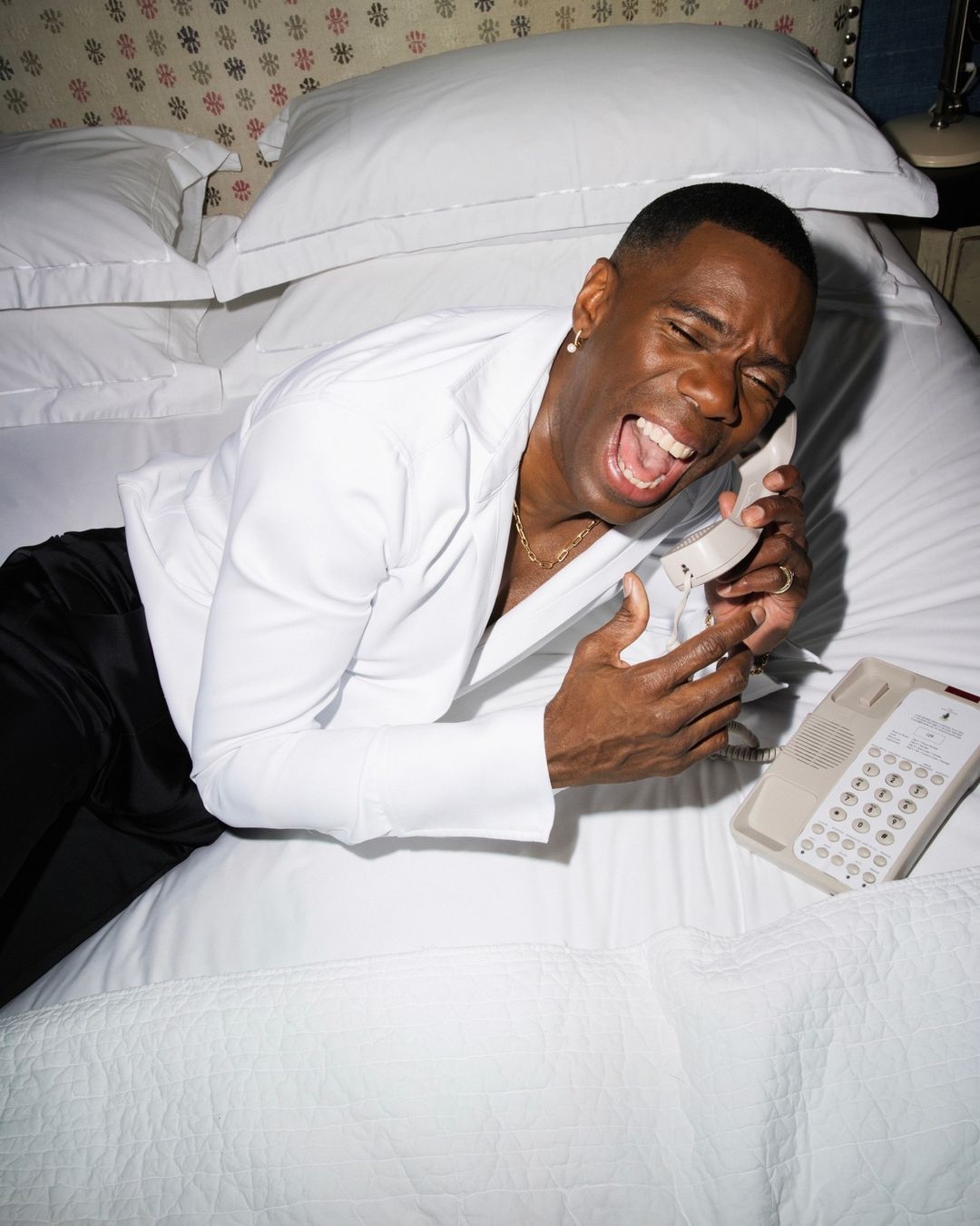 Adult male in a white shirt and black pants lies on a white bed while holding a landline telephone with an open mouth and closed eyes.