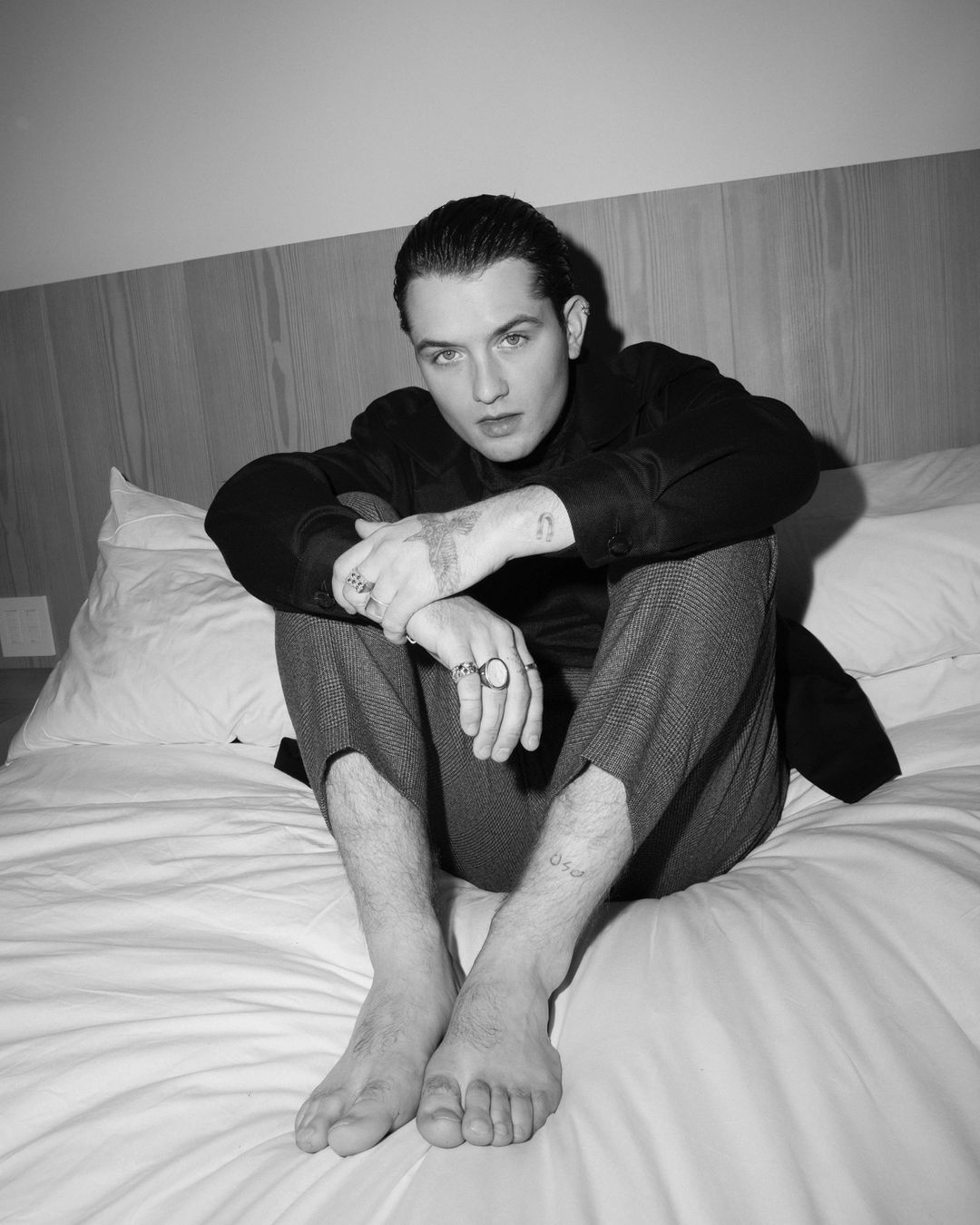 Monochrome portrait of a young man sitting barefoot on a white hotel bed with dark trousers and a black shirt.