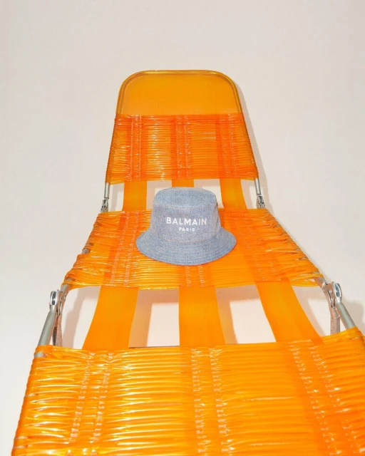 Denim bucket hat by Balmain Paris on a bright orange vintage lawn chair. The scene is lit with a soft, studio light.