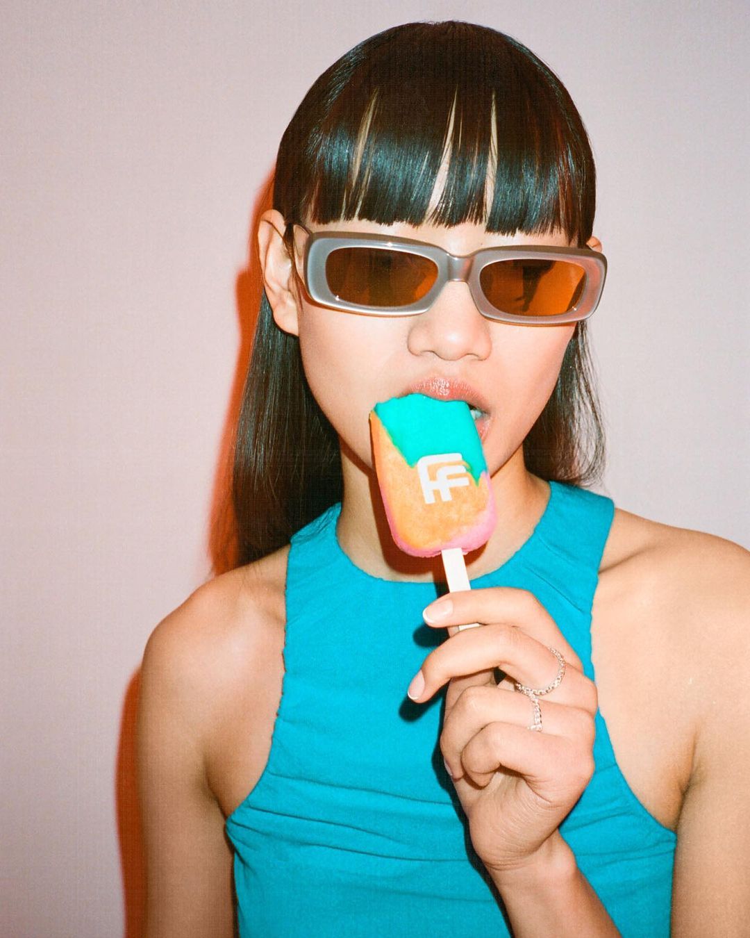 Young adult woman with straight dark hair and sunglasses eating a colorful popsicle indoors with a light background.