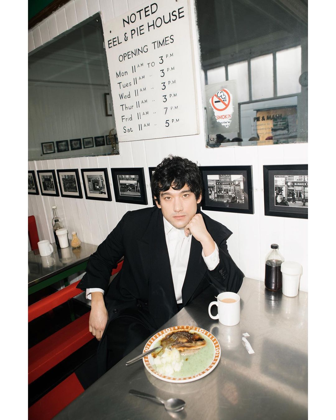 Man in a suit sitting at a diner table with a plate of food and a mug in an indoor diner with red and black accents and natural light.