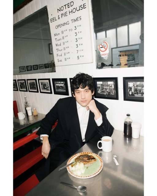 Man in a suit sitting at a diner table with a plate of food and a mug in an indoor diner with red and black accents and natural light.