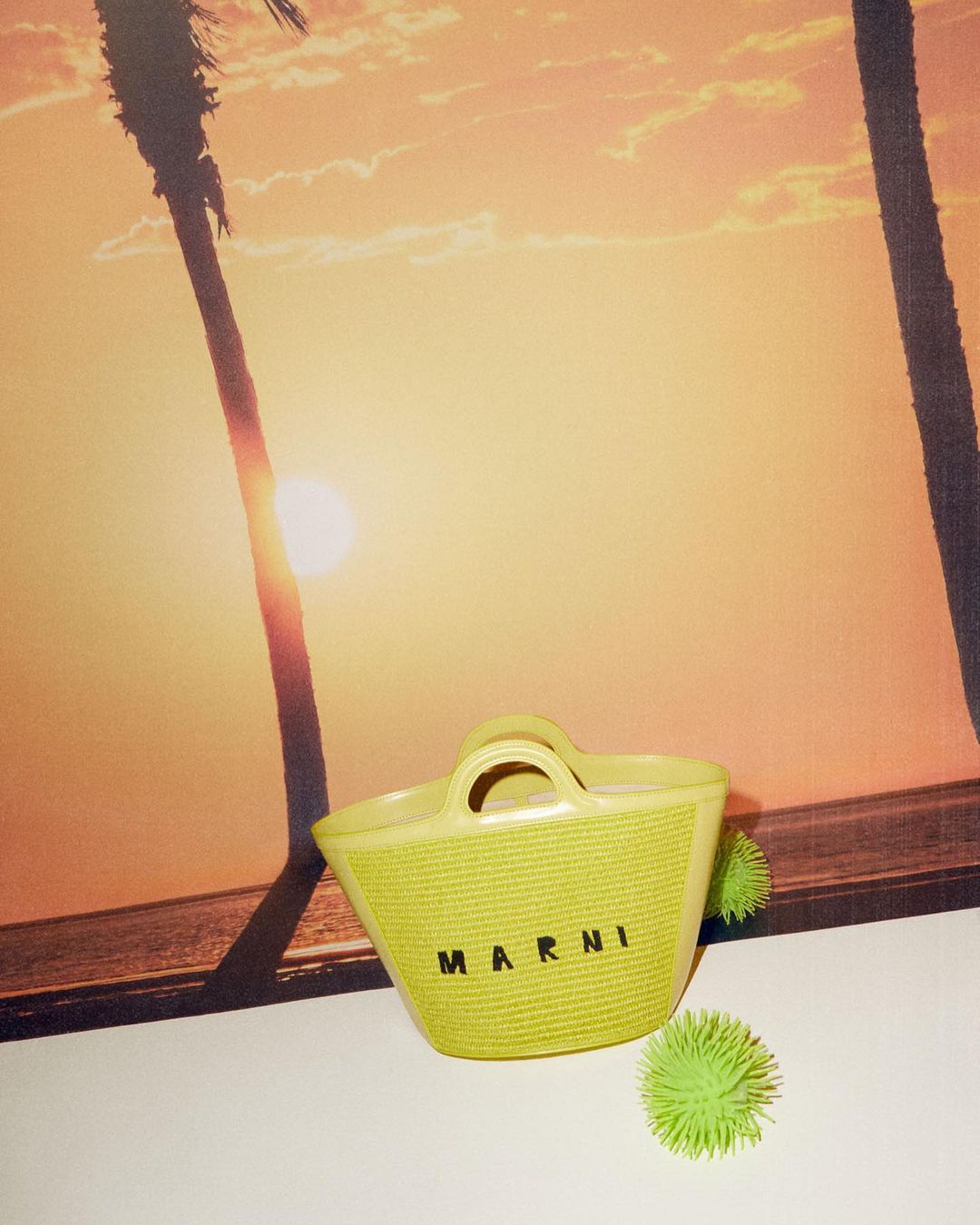 Yellow woven tote bag with 'MARNI' text sits beside spiky green balls, set against a backdrop of silhouetted palm trees at sunset.