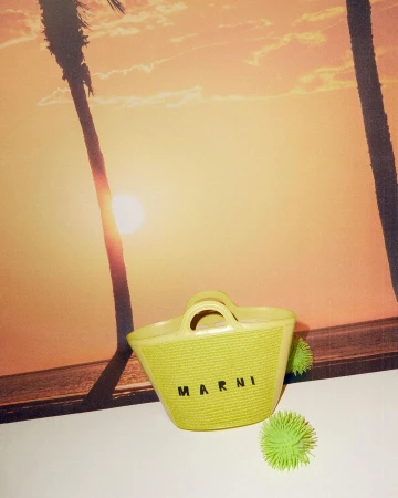 Yellow woven tote bag with 'MARNI' text sits beside spiky green balls, set against a backdrop of silhouetted palm trees at sunset.