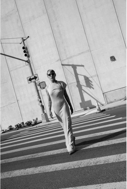 A person in a dress walks across a crosswalk in an urban setting, with a traffic light and a large building in the background.