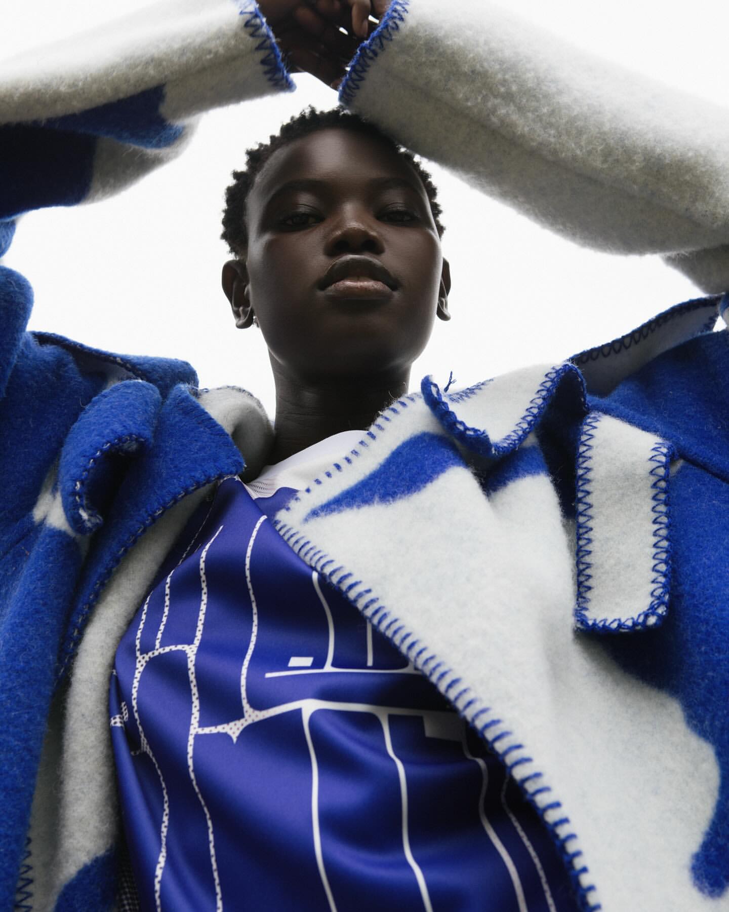 Portrait of a young Black woman in a patterned jacket looking at the camera from a low angle with a white background.