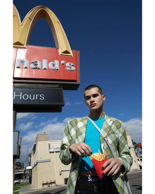 Young adult male model stands outside a McDonald's holding French fries, wearing a patterned blazer, blue sweater, and black pants. 