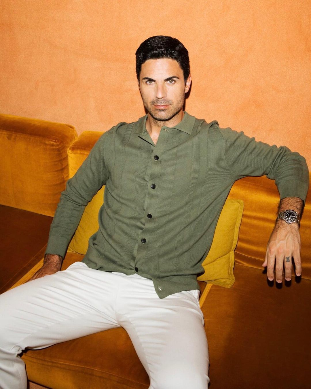 Man sitting on a yellow velvet sofa, wearing a green button-down shirt and white pants, posing in a casual indoor setting.