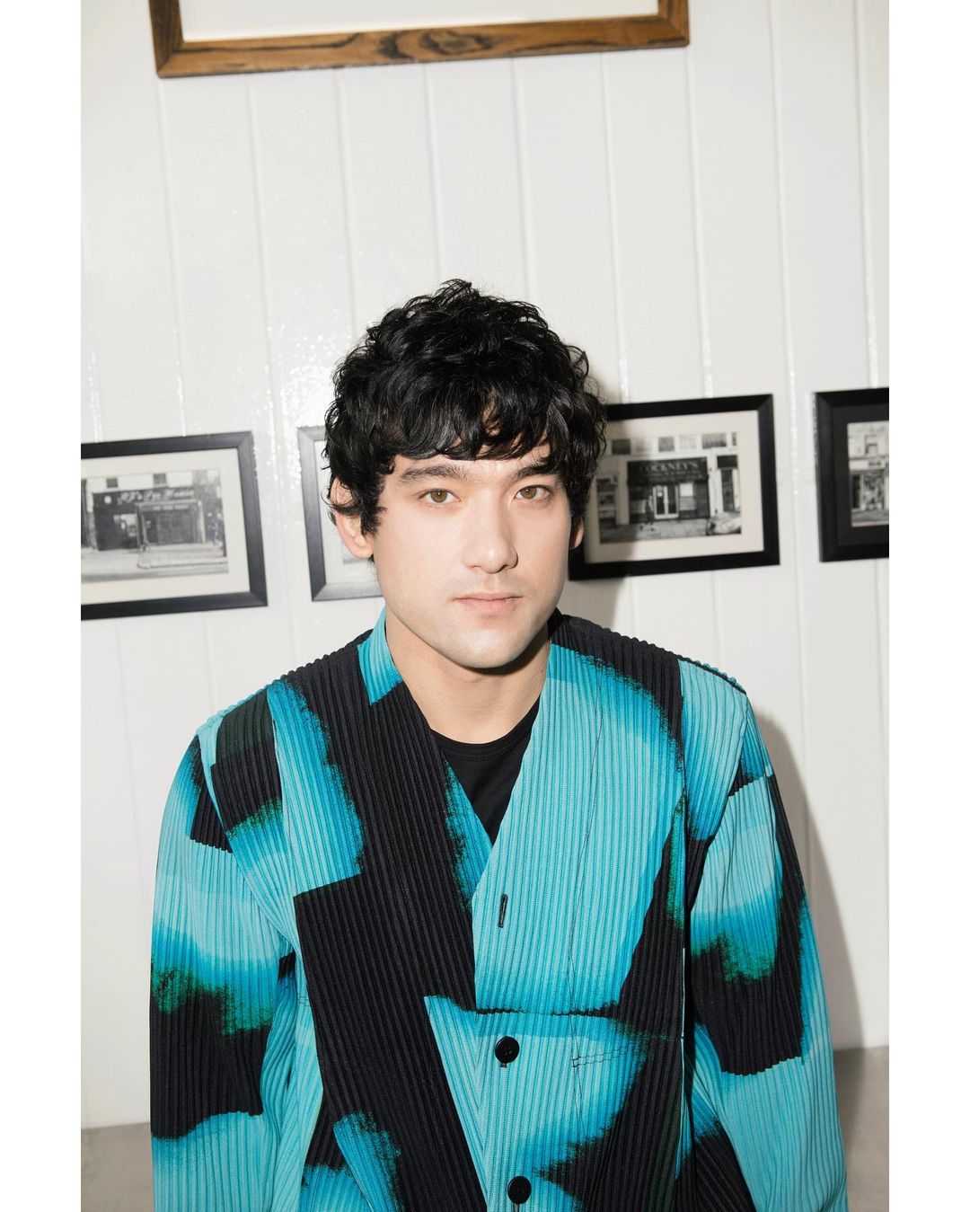 Portrait of a young adult male with dark curly hair wearing a patterned blue and black jacket, posing indoors against a white paneled wall.