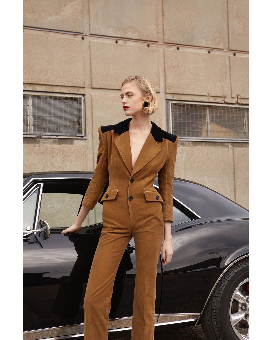 A woman in a brown corduroy suit stands by a classic black car, posing in front of a beige building on an overcast day.