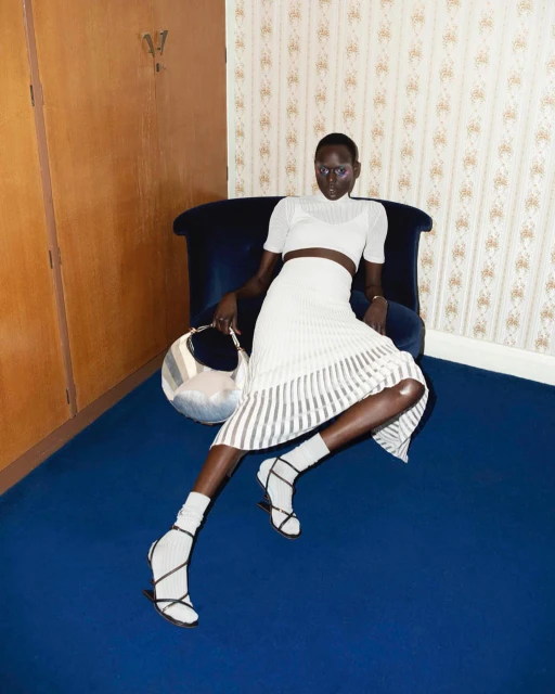 Fashion shot of a Black female model wearing a white knit outfit and strappy heels, seated on a blue chair in an indoor studio.